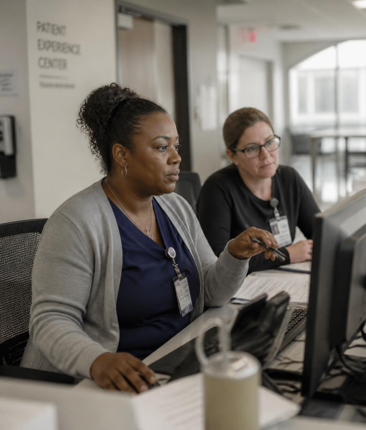 Two healthcare administrators working side by side at a shared workstation in a daylit hospital corridor